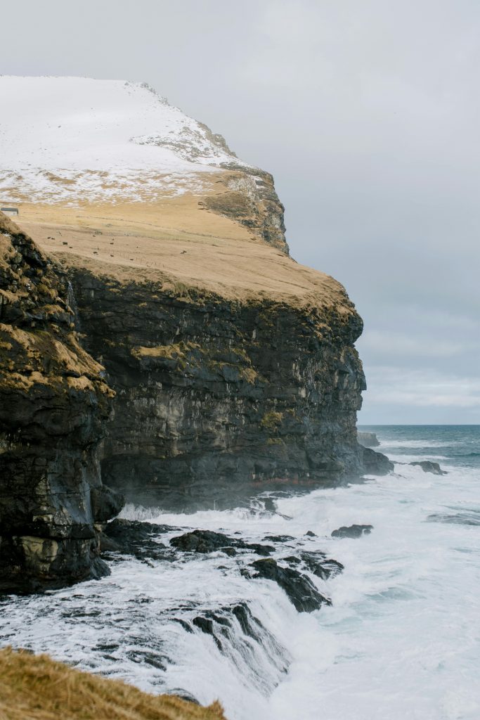 Home Dramatic cliffs on the Faroe Islands coastline with waves crashing against the shore under a cloudy sky.